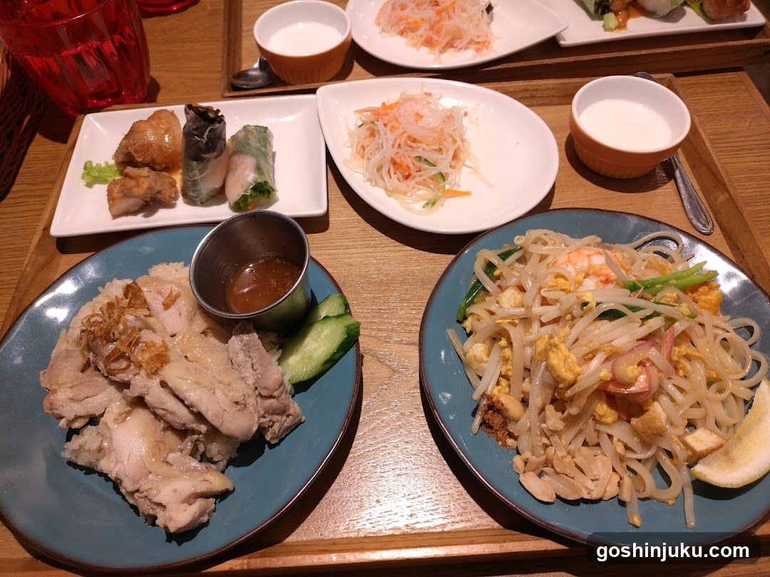 A wooden table displays a variety of Asian dishes including stir-fried noodles with shrimp, sliced chicken with cucumber, spring rolls, shredded vegetables, and small bowls of sauce or soup.