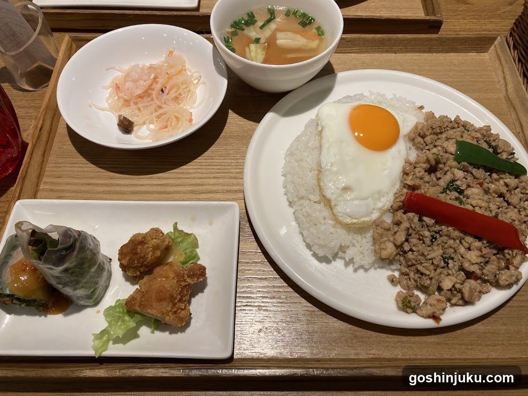 A wooden tray holds a variety of dishes including rice topped with a sunny-side-up egg, minced pork mixed with chili peppers, spring rolls, fried chicken pieces, shredded vegetables in sauce, and a bowl of soup with tofu and green onions.