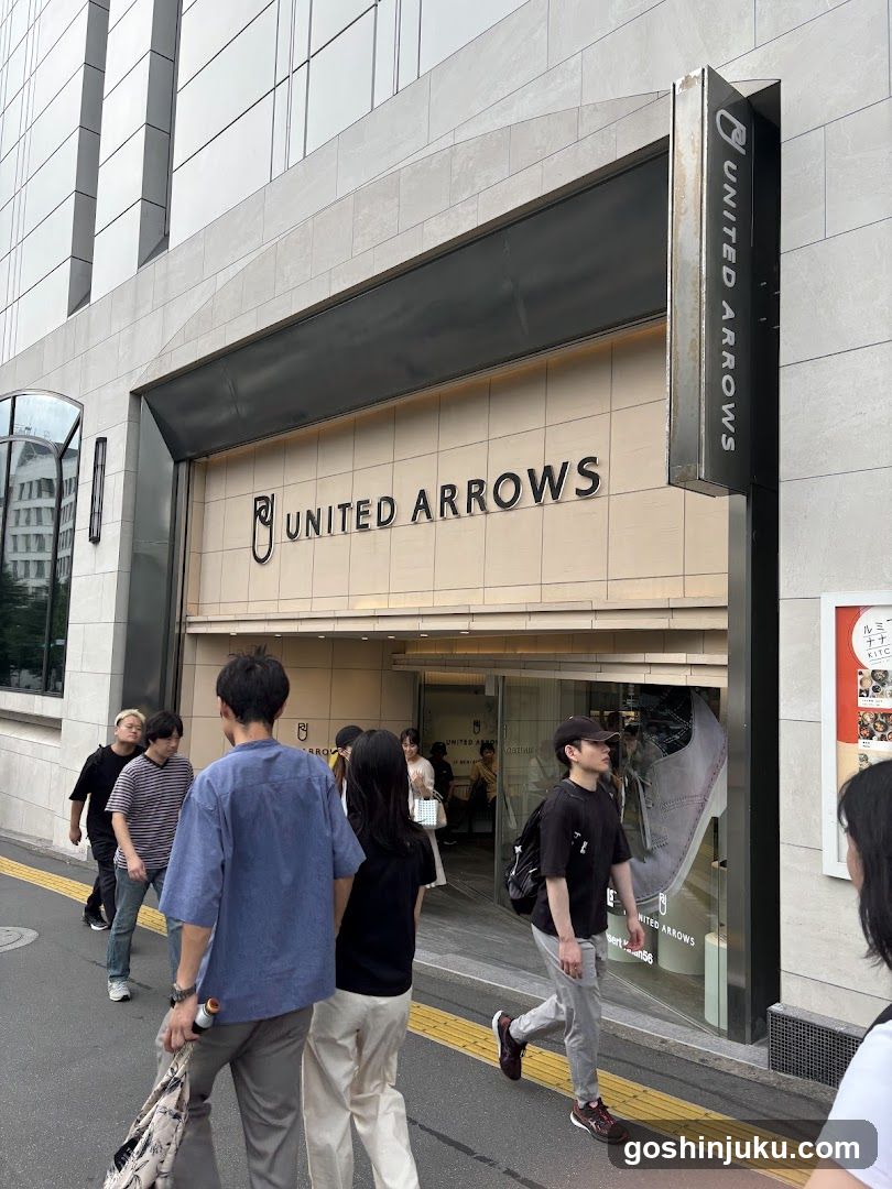 Photo of the exterior entrance and storefront facade of a United Arrows store. The building features modern architecture with large glass windows and beige cladding. Prominent signage displays 'UNITED ARROWS' on the facade and vertically along the side column. Pedestrians are walking past the shop, including individuals in casual attire such as t-shirts and jeans. A poster is visible to the right of the entrance.