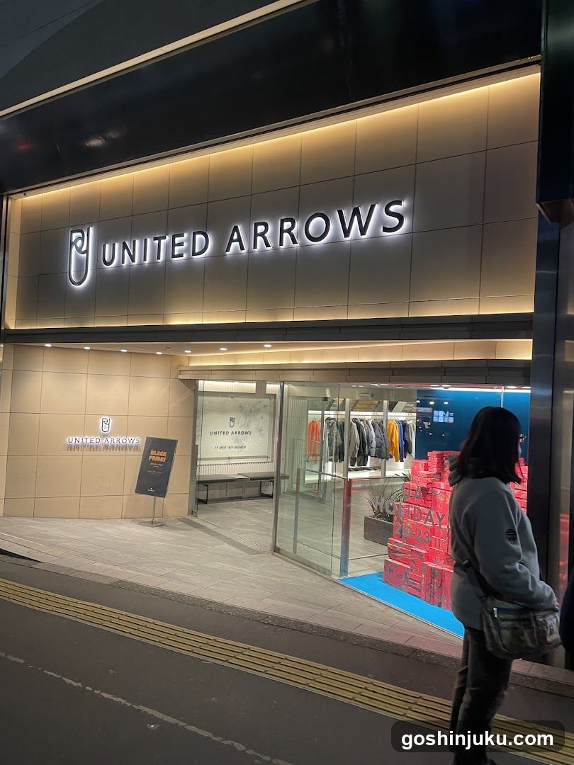 A modern storefront with illuminated 'UNITED ARROWS' signage above the entrance. The glass doors are open, revealing racks of clothing inside. A person stands near the entrance on the sidewalk.