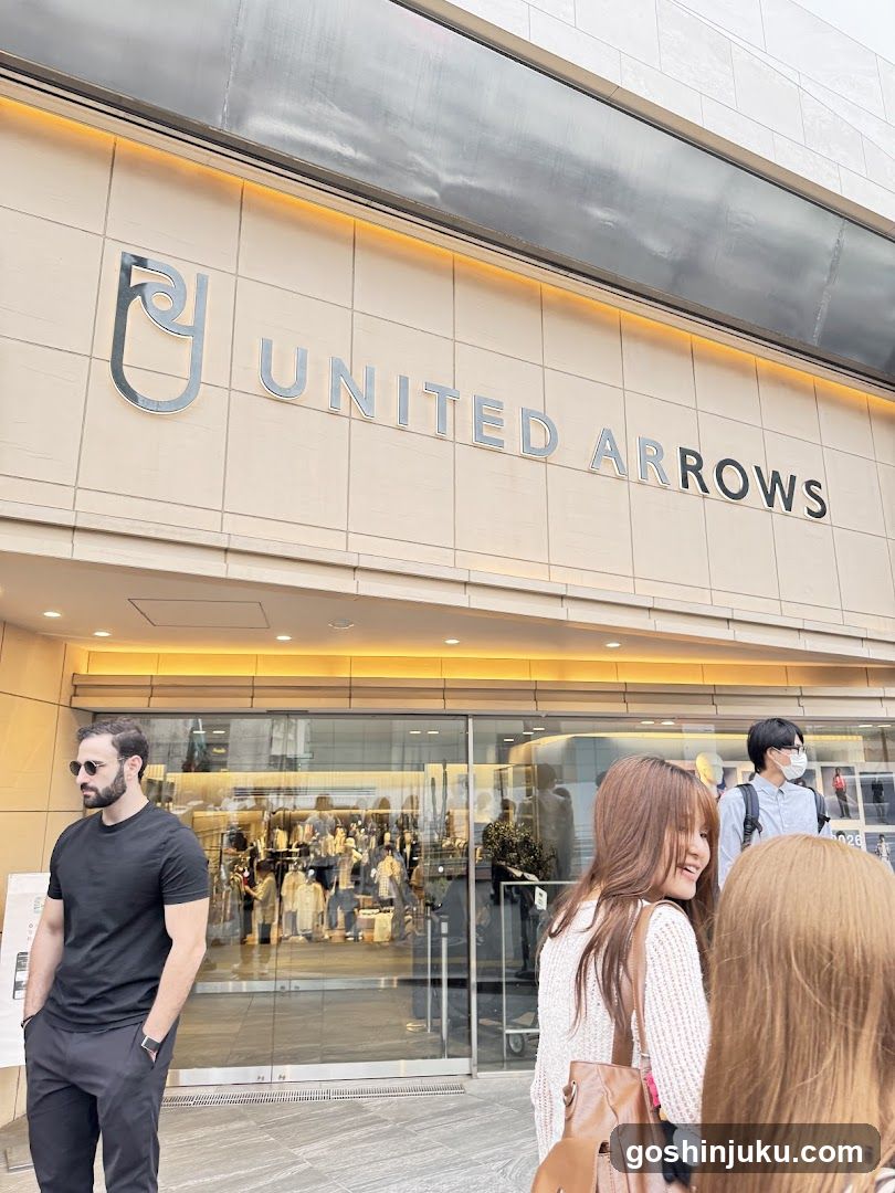 Photo of the exterior entrance and storefront facade of a United Arrows store, featuring prominent signage with the brand logo and name mounted on the building's beige tiled wall above glass doors.