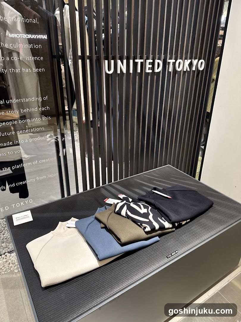 A photo of the United Tokyo store entrance, featuring a dark slatted facade with 'UNITED TOKYO' signage in white letters. A glass window displays partial text about craftsmanship and creativity. In front, a black textured display table holds neatly folded clothing items: cream, blue, olive green, patterned, and navy garments.