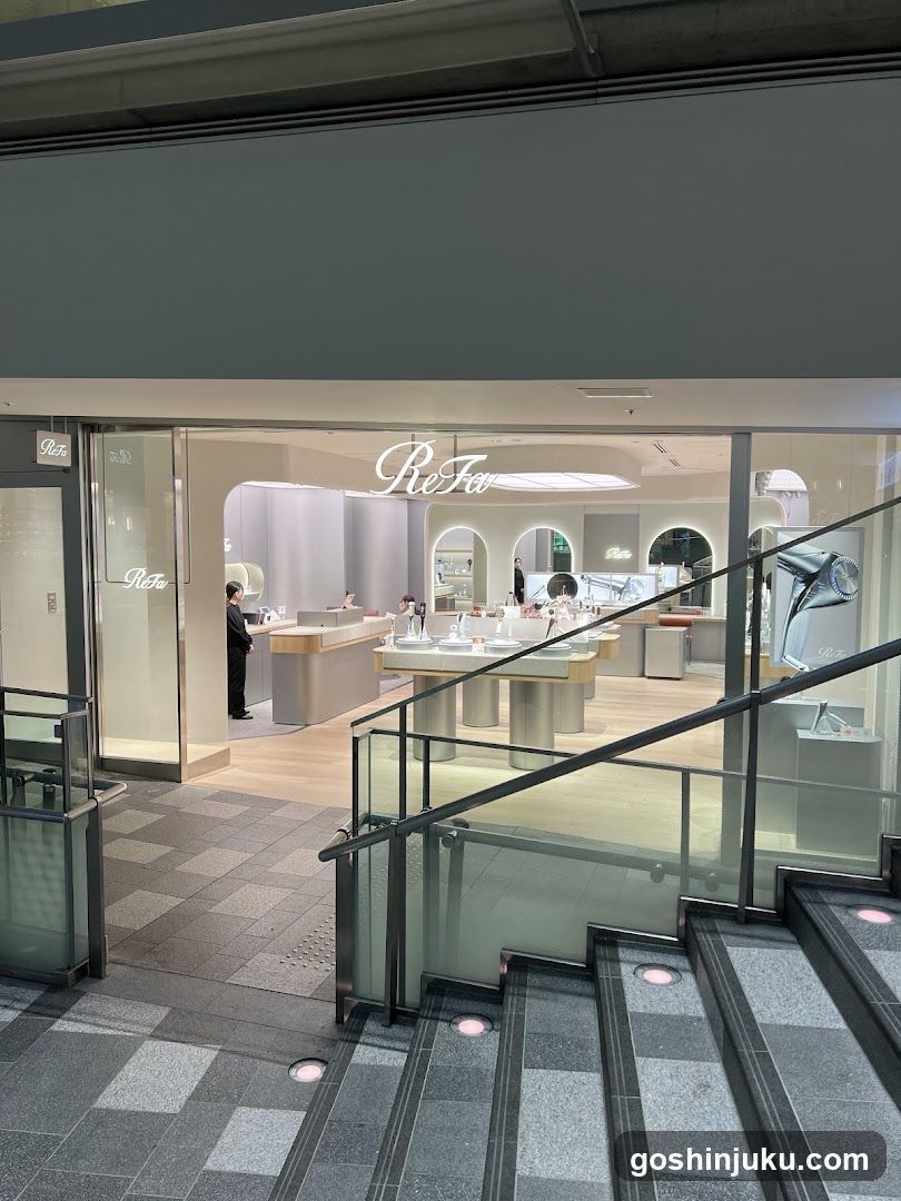 A modern retail store with glass facade and visible signage reading 'Re-Fit' above the entrance. Interior displays include product tables, staff members, and decorative elements like a large metallic sculpture. The storefront is located in a mall setting with tiled flooring and staircases nearby.