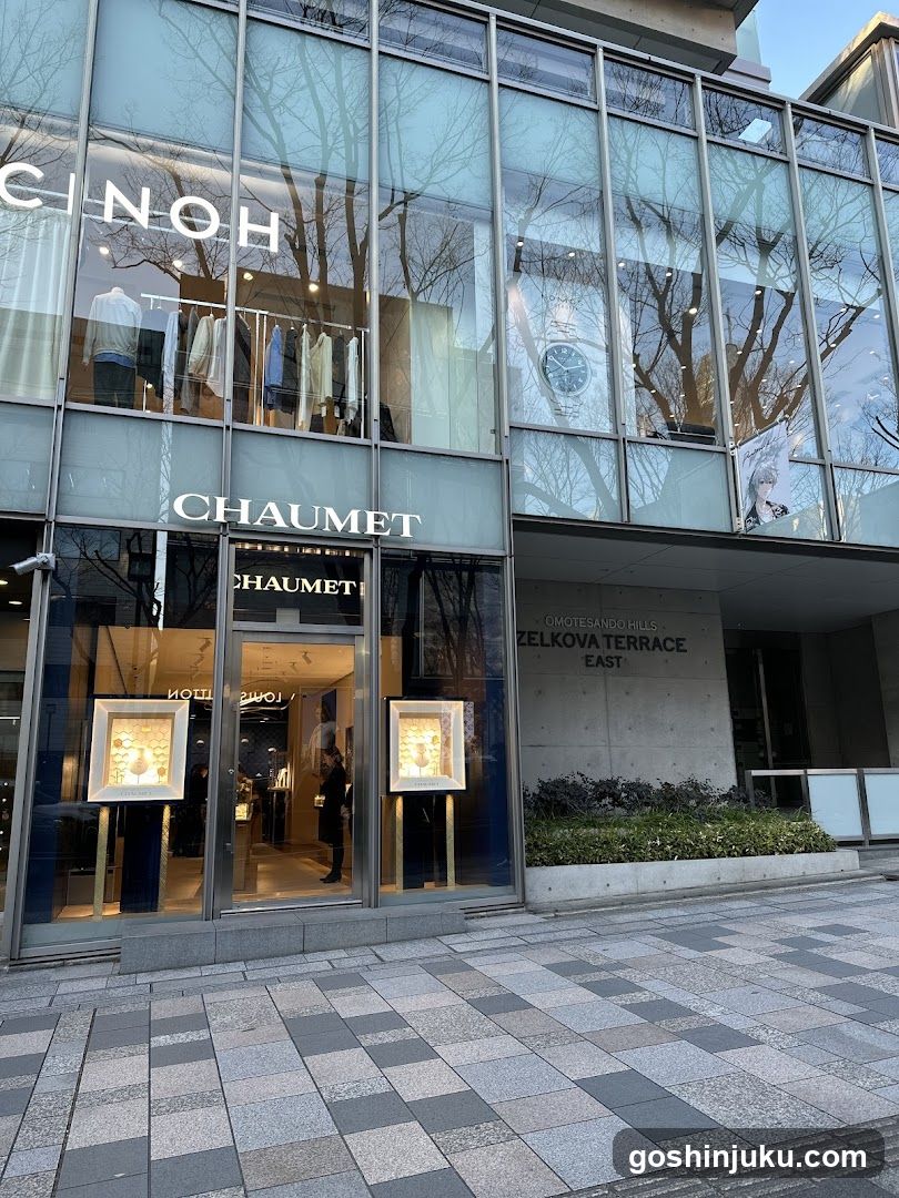 A modern glass-fronted shop with 'CHAUMET' signage prominently displayed above the entrance and on the upper facade. The store features large windows showcasing jewelry displays, including a watch visible inside. A person is seen entering through the automatic doors. To the right of the entrance, a sign reads 'OMOTE SANO MILES ZELKOVA TERRACE EAST'. The building exterior includes reflective glass panels showing tree reflections.