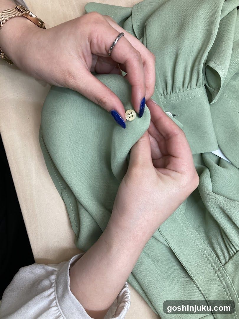 Close-up of two hands with blue nail polish carefully positioning a small cream-colored button on the sleeve of a light sage-green blouse or dress. The fabric appears soft and slightly wrinkled, suggesting it is being prepared or fitted. A gold bracelet is visible on one wrist, and another hand wears a thin silver ring. The background includes part of a wooden surface and hints of white ruffled cuffs.