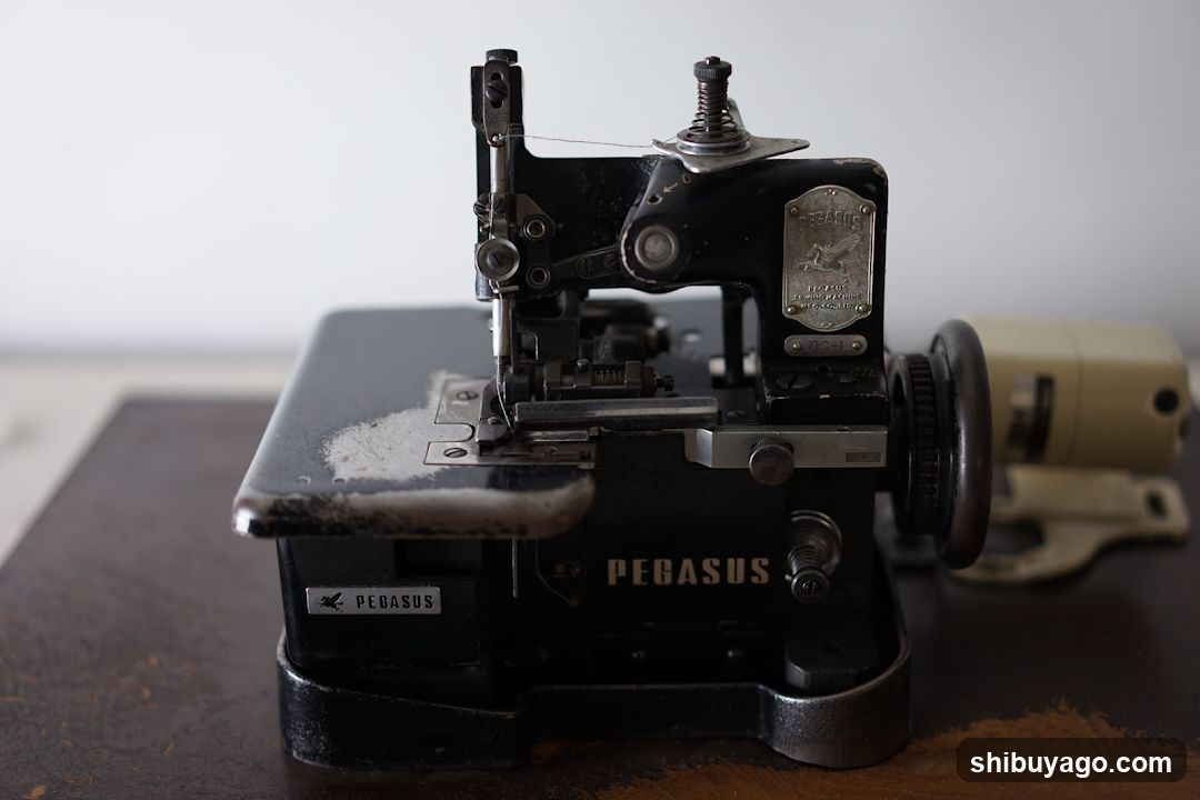 A close-up of a black vintage sewing machine with the brand name 'Pegasus' visible on its side and top plate. The machine features intricate mechanical components, including a presser foot, needle bar, and handwheel. A white powdery substance is present on the flat bed near the needle area. In the background, part of another beige-colored device is partially visible.
